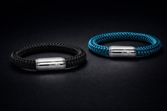 Zwei hochwertige Segeltauarmbänder in Schwarz und Türkis liegen leicht versetzt auf dunklem, mattem Untergrund. Beide Armbänder besitzen einen polierten Edelstahl-Magnetverschluss mit graviertem Dreizack-Symbol. Luxuriöse Still-Life-Produktfotografie mit präziser Studioausleuchtung, klaren Konturen und edler Premium-Ästhetik.