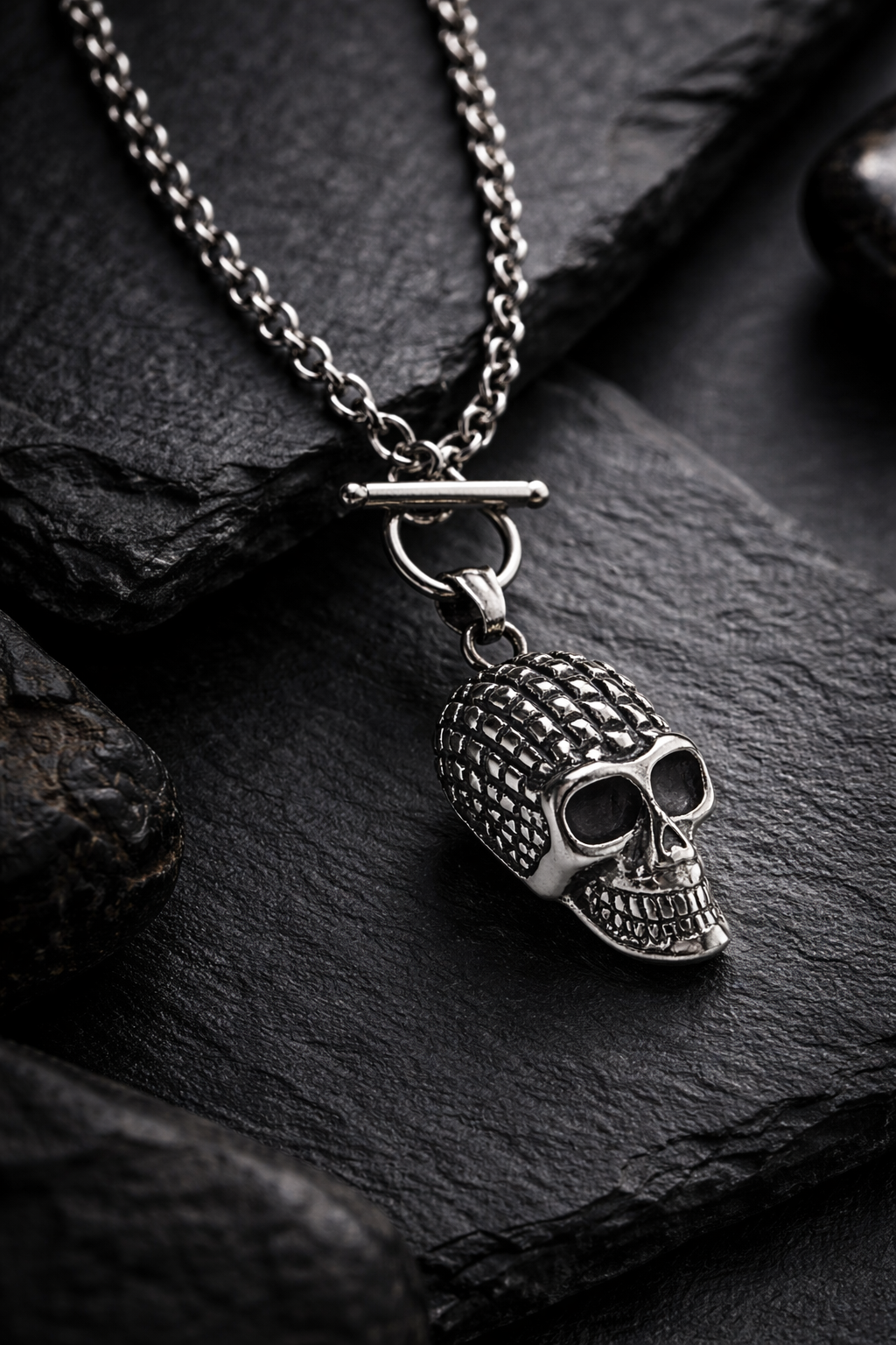 Silberfarbene Totenkopf-Halskette aus Edelstahl mit Gliederkette, als hochwertige Still-Life-Produktfotografie in dunkler, luxuriöser Premium-Ästhetik auf schwarzem Steinuntergrund.