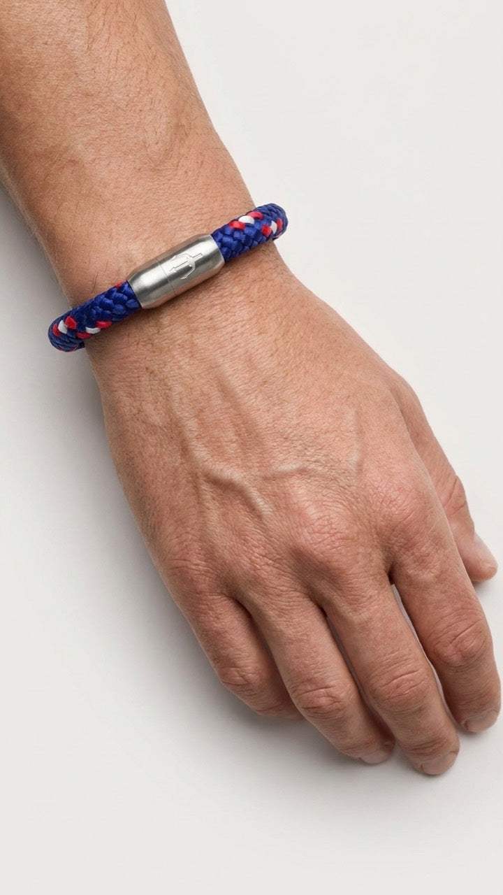 Blau-rot-weisses geflochtenes Armband mit silberfarbenem Dreizack Verschluss am Handgelenk vor hellem Studiohintergrund.