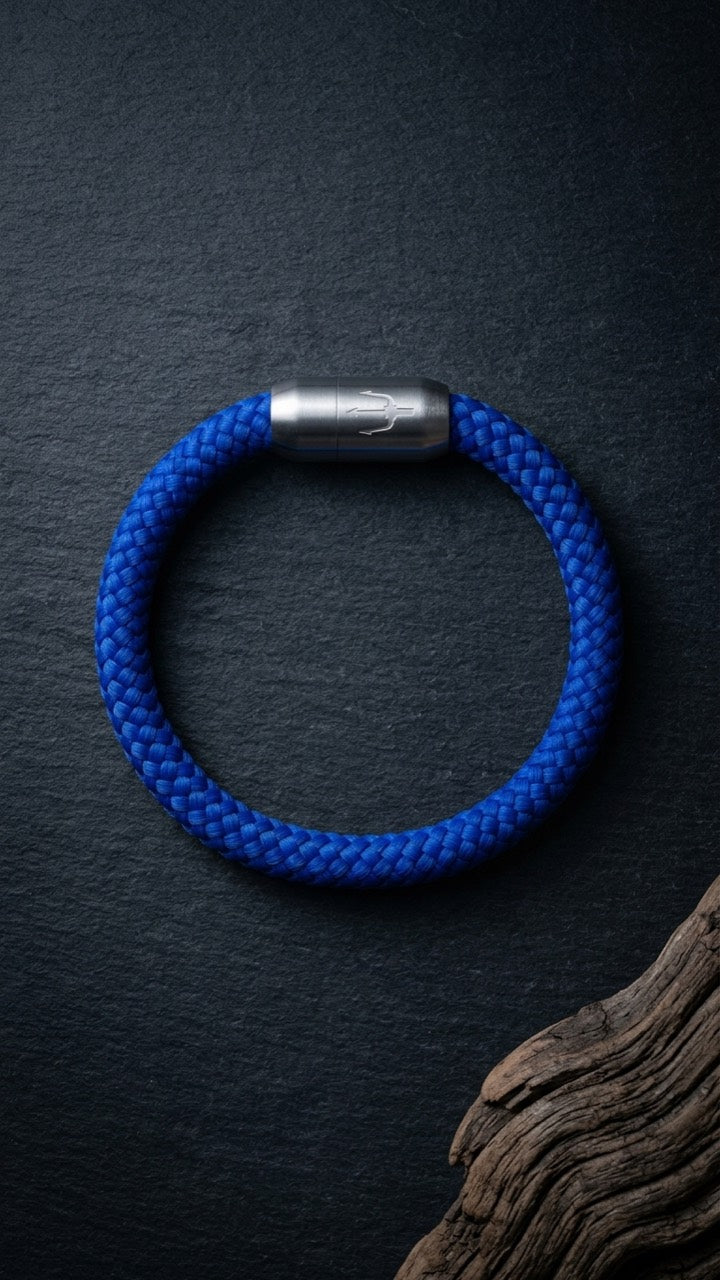Blaues geflochtenes Armband mit silberfarbenem Dreizack Verschluss als Produktfoto auf dunklem Schiefer mit Holzdetail.