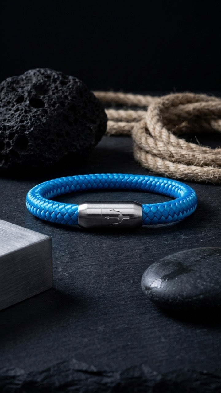 Blaues geflochtenes Armband mit silberfarbenem Dreizack Verschluss als Stillleben mit Stein, Seil und Metallblock.