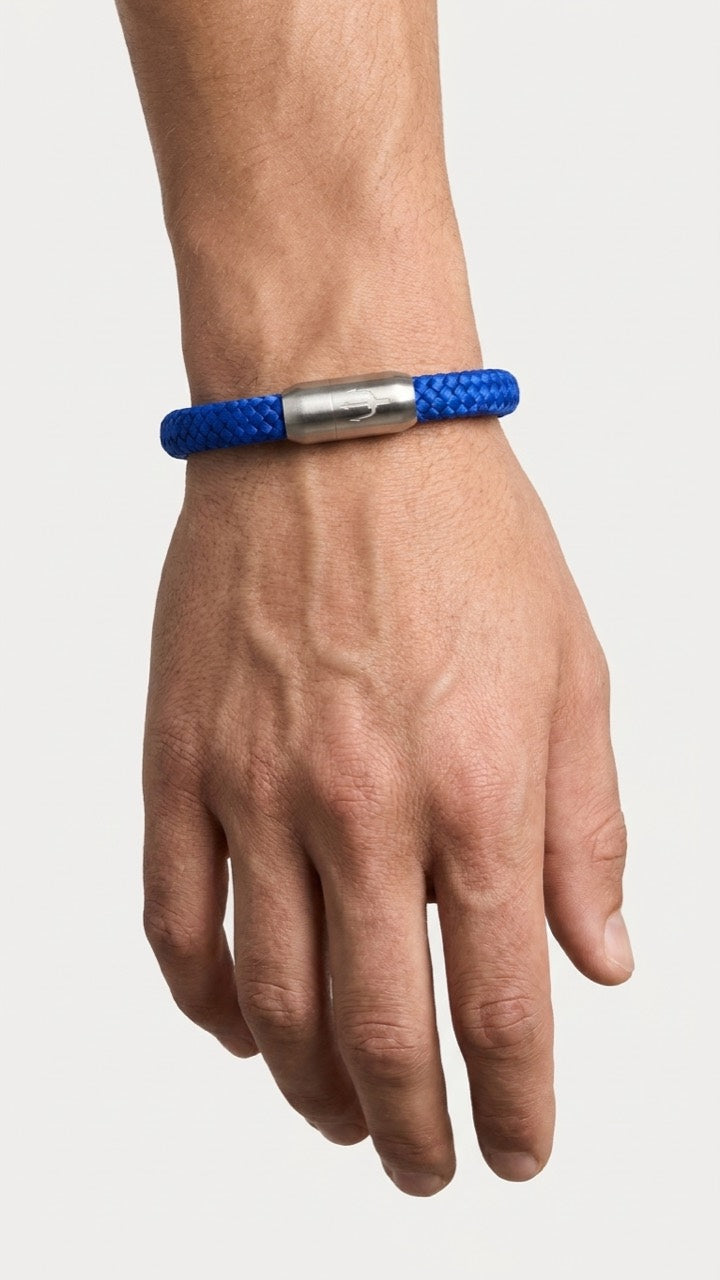 Blaues geflochtenes Armband mit silberfarbenem Dreizack Verschluss am Handgelenk vor hellem Studiohintergrund.