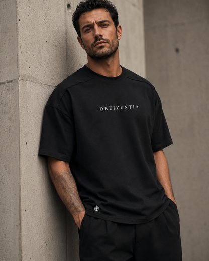 Model in schwarzem DREIZENTIA Essential Raw Herren-T-Shirt lehnt an einer Betonwand