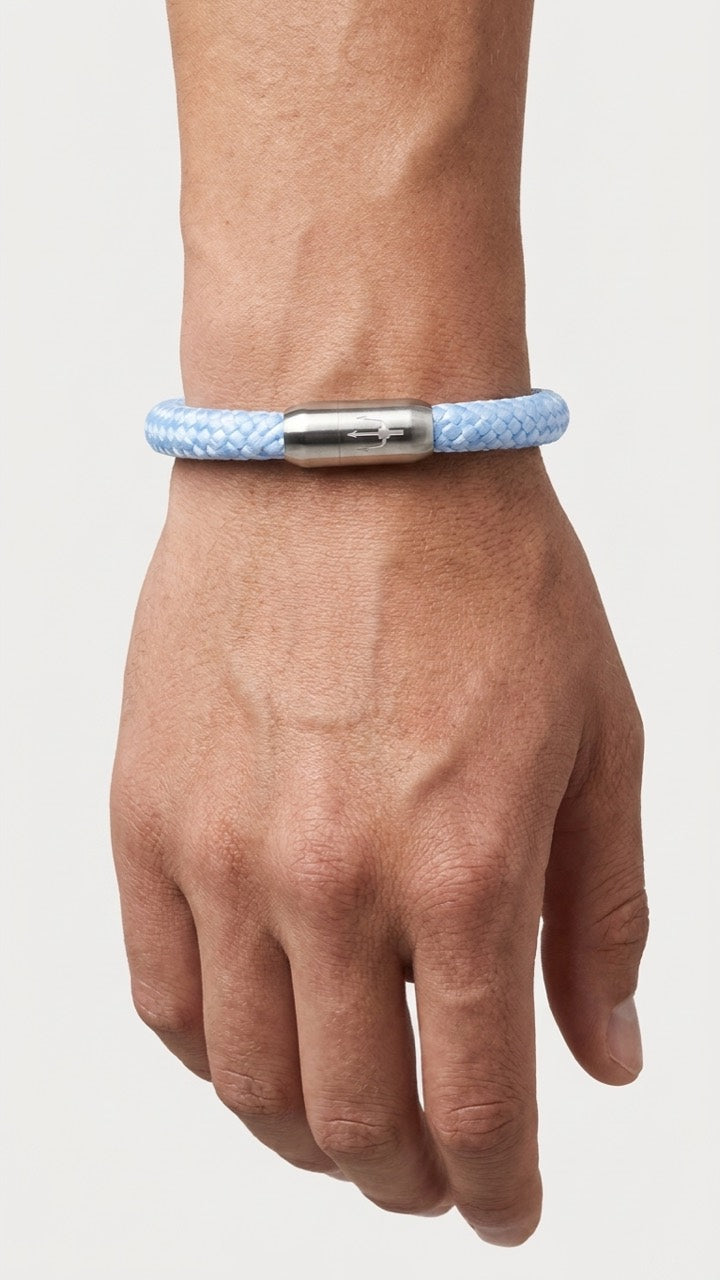 Hellblaues geflochtenes Armband mit silberfarbenem Dreizack Verschluss am Handgelenk vor hellem Studiohintergrund.