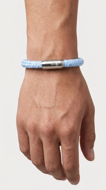 Hellblaues geflochtenes Armband mit silberfarbenem Dreizack Verschluss am Handgelenk vor hellem Studiohintergrund.