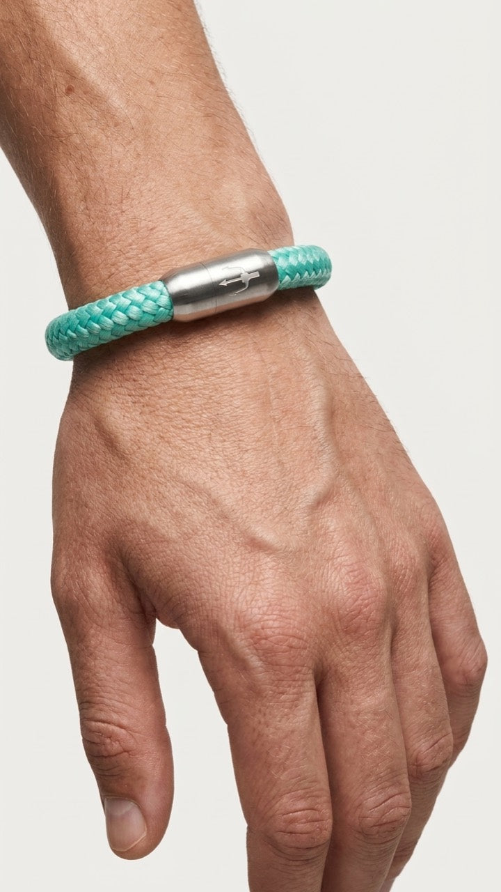 Mintfarbenes geflochtenes Armband mit silberfarbenem Dreizack Verschluss am Handgelenk vor hellem Studiohintergrund.
