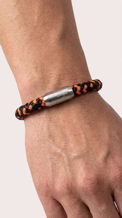 Schwarz-oranges geflochtenes Armband mit silberfarbenem Dreizack Verschluss am Handgelenk vor hellem Studiohintergrund.