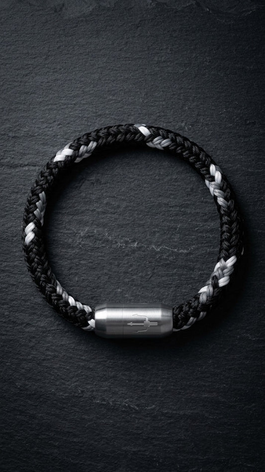 Schwarz-weisses geflochtenes Armband mit silberfarbenem Dreizack Verschluss als Produktfoto auf dunklem Schiefer.