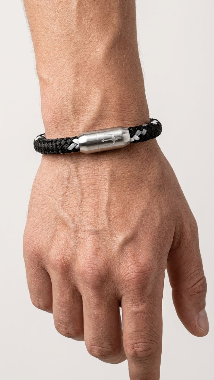 Schwarz-weisses geflochtenes Armband mit silberfarbenem Dreizack Verschluss am Handgelenk vor hellem Studiohintergrund.