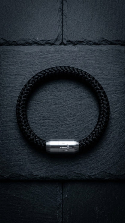 Schwarzes geflochtenes Armband mit silberfarbenem Dreizack Verschluss als Produktfoto auf dunklem Schiefer.