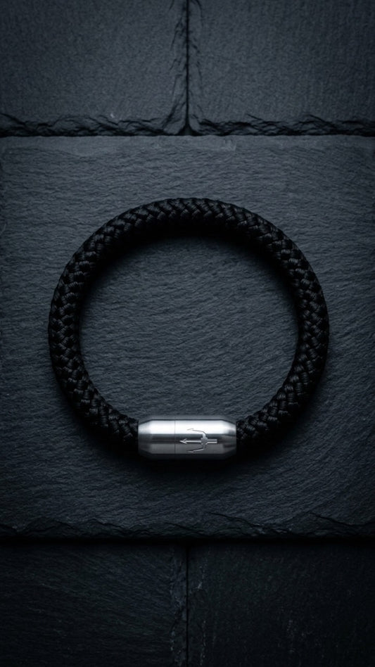 Schwarzes geflochtenes Armband mit silberfarbenem Dreizack Verschluss als Produktfoto auf dunklem Schiefer.