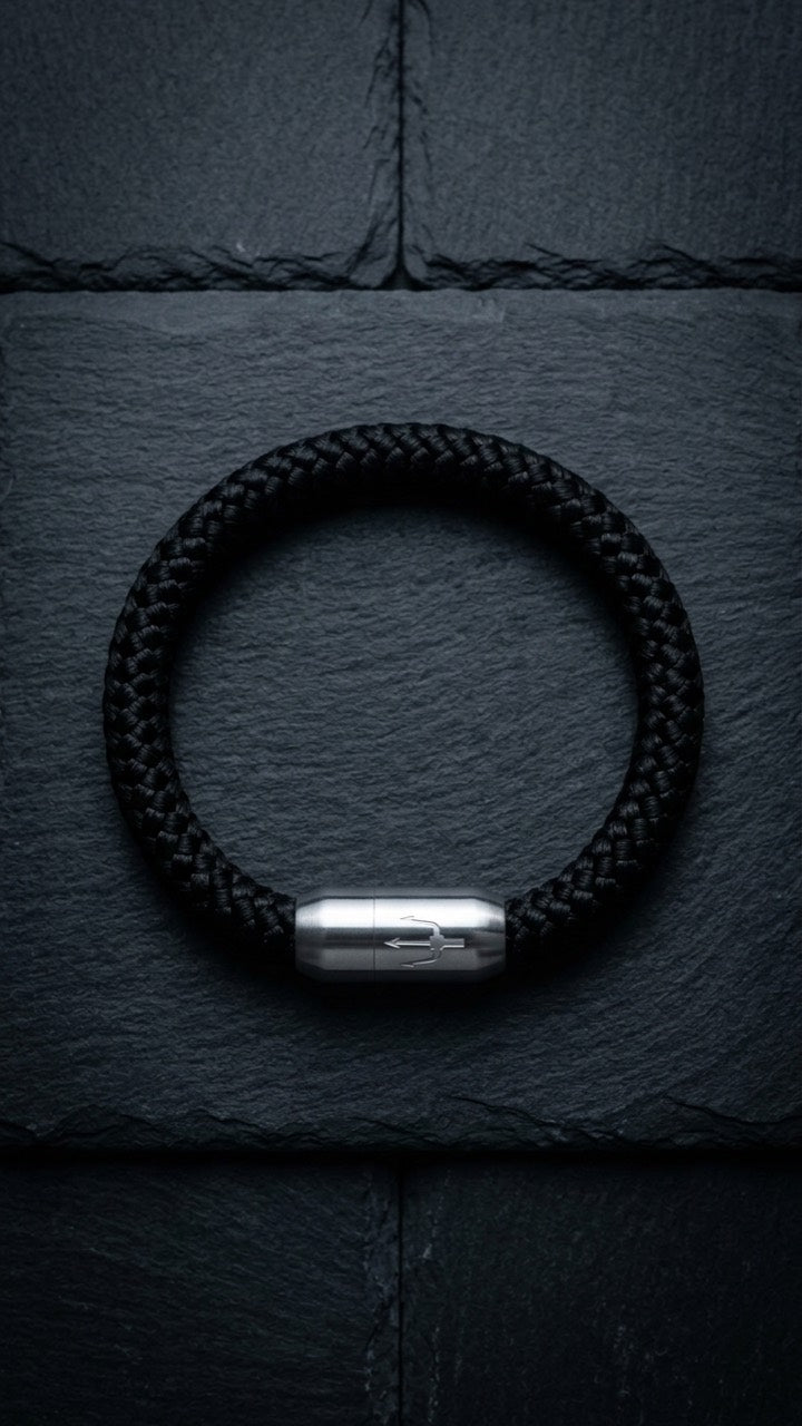 Schwarzes geflochtenes Armband mit silberfarbenem Dreizack Verschluss als Produktfoto auf dunklem Schiefer.