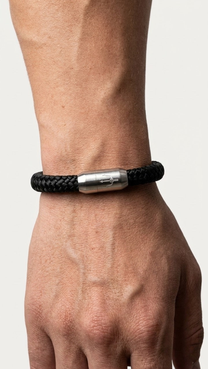 Schwarzes geflochtenes Armband mit silberfarbenem Dreizack Verschluss am Handgelenk vor hellem Studiohintergrund.