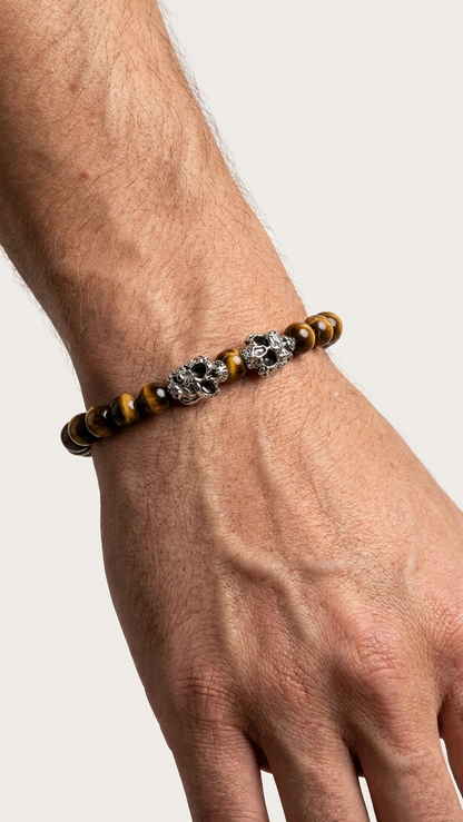 Tigerauge-Perlenarmband mit zwei silberfarbenen Schaedelperlen am Handgelenk vor hellem Studiohintergrund.