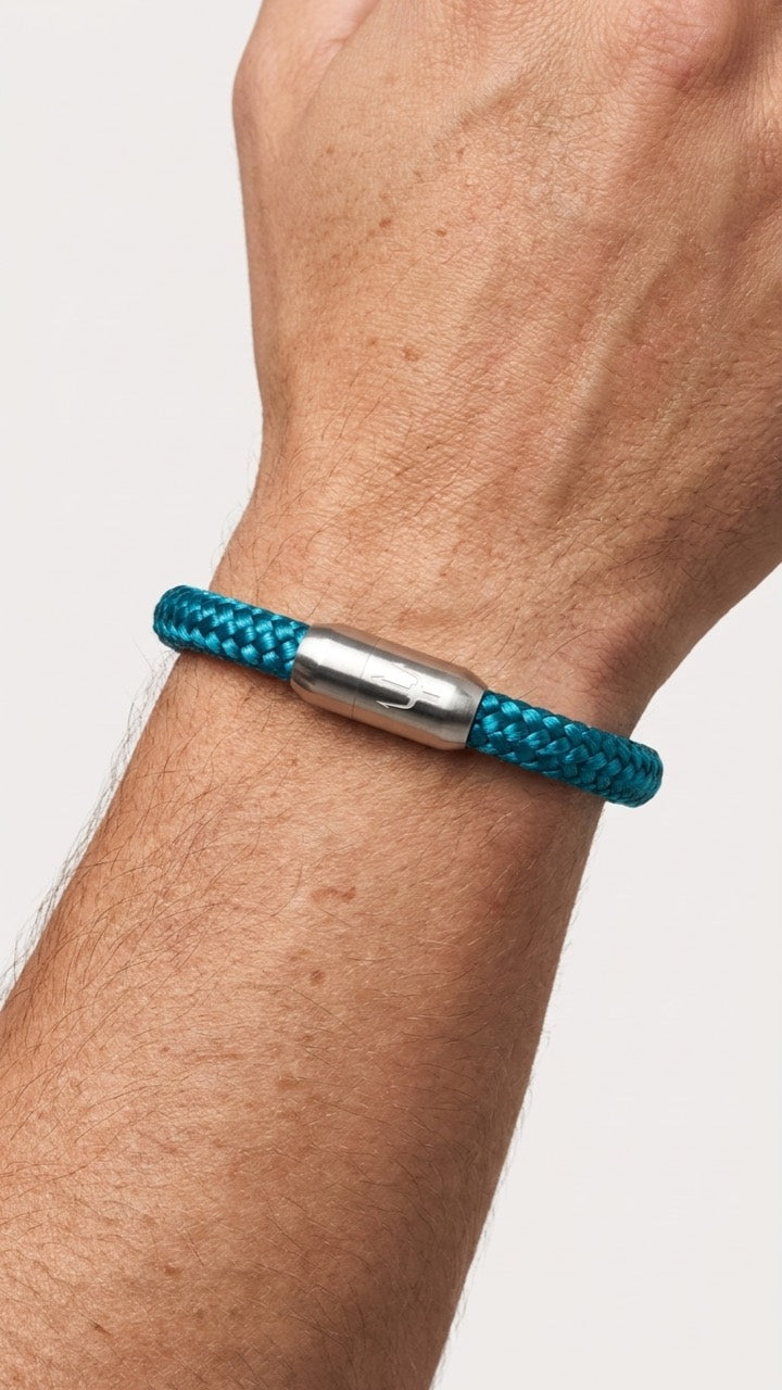 Tuerkisfarbenes geflochtenes Armband mit silberfarbenem Dreizack Verschluss am Handgelenk vor hellem Studiohintergrund.
