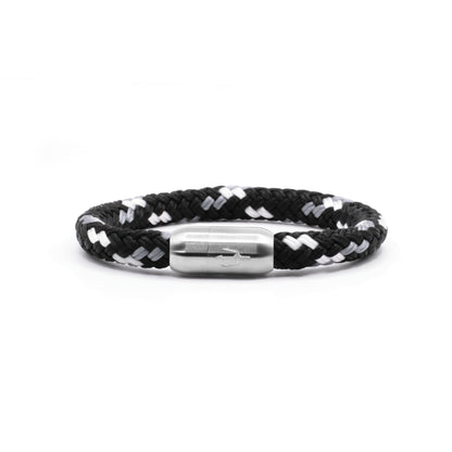 “Geflochtenes Armband in Schwarz-Weiß mit einem Edelstahl-Magnetverschluss, verziert durch eine eingravierte Dreizack-Symbolik. Modernes und minimalistisches Design, inspiriert von Wellen und Kontrasten.”